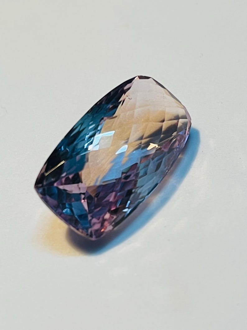 May include: A rectangular-cut gemstone displaying a colour gradient, shifting from deep blue to purple and fading into a pale pink. The facets of the stone reflect light, producing a sparkling effect. This gemstone is suitable for jewellery making.