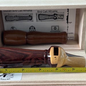 May include: A wooden bird call whistle, with a dark reddish-brown body and a lighter wood accent. The whistle is displayed in a wooden box with a diagram. A measuring tape shows the whistle is approximately 6 inches long.