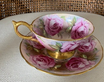 Può includere: Un set vintage di tazza da tè e piattino con un delicato motivo floreale. La tazza e il piattino sono decorati con rose rosa e foglie verdi su sfondo bianco. La tazza ha un bordo dorato e un manico dorato.