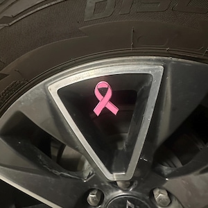 May include: A pink ribbon emblem, a symbol of breast cancer awareness, is attached to the centre of a vehicle's wheel. The wheel is dark grey with a silver trim, and the tyre is black.