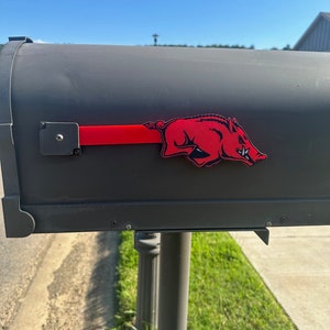 May include: A black mailbox with a red flag and a red Razorback logo. The Razorback logo is a stylized image of a running wild boar. The mailbox is mounted on a gray post.