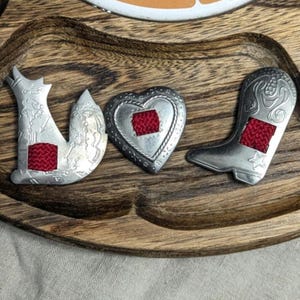 May include: Silver-toned metal pins spelling "LOVE" with a fox, heart, and cowboy boot design. Each pin features a square of red woven fabric. The pins are displayed on a wooden tray with a natural wood grain.