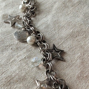 May include: A silver-toned charm bracelet featuring multiple dangling elements including white faux pearls, clear faceted beads, solid stars, open-frame stars, and a round charm engraved with a crescent moon and small star symbols.