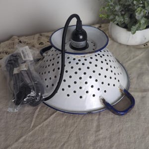 May include: A white colander repurposed as a pendant light fixture. The colander has a blue rim and handles, with a black electrical cord and bulb socket. A package of black electrical cord is also visible.