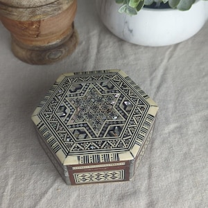 Puede incluir: Una caja de joyería hexagonal decorativa con un diseño de estrella en la tapa. La caja presenta incrustaciones de nácar y patrones geométricos en tonos crema, marrón y negro. La caja mide aproximadamente 10 cm de ancho y 5 cm de alto.