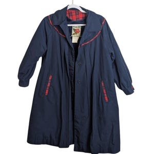 May include: A navy blue coat featuring a red and black plaid lining, a collar with plaid trim, and matching plaid accents on the pocket openings. The coat has long sleeves with cuffs, a button-front closure, and a label with a rose graphic inside.