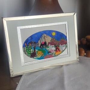 May include: A rectangular silver-toned frame containing an oval-shaped textile art piece. The embroidery depicts a landscape with mountains, a yellow sun, white houses, cacti, and animals including a llama, all rendered in vibrant thread colors.