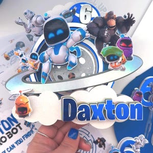 Puede incluir: Un adorno para tarta de cumpleaños con temática de robots. El adorno incluye un personaje de robot, un gorila y otros personajes. El nombre "Daxton" se muestra en letras azules. El número "6" también es visible.