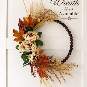 May include: A circular wreath featuring dark brown wooden beads on one side and a cluster of cream-colored peonies, orange and red maple leaves, and dried grasses on the other. The text "Matching Wreath Also Available!" is printed in the upper right.