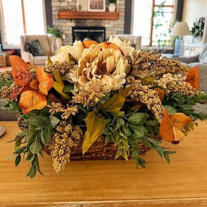 May include: A floral arrangement in a woven basket featuring cream-colored peonies, orange autumn leaves, and textured golden-brown sprigs. The arrangement is displayed on a wooden surface next to a white pillar candle in a decorative holder.