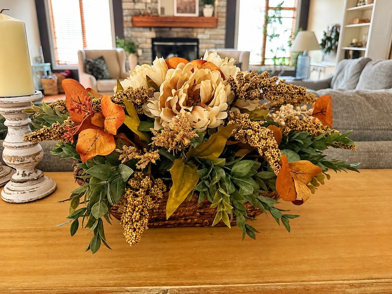 May include: A floral arrangement in a woven basket featuring cream-colored peonies, orange autumn leaves, and textured golden-brown sprigs. The arrangement is displayed on a wooden surface next to a white pillar candle in a decorative holder.