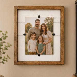 May include: A rectangular wall-mounted photo frame featuring a dark brown stained wooden border and a white horizontal plank background. A portrait of a family of four is secured to the center with four small black metal clips at the edges.