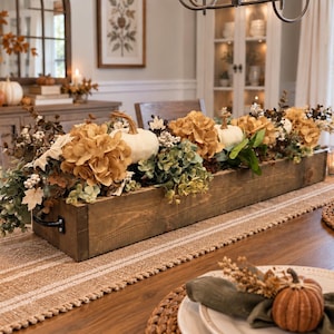 May include: A long, rectangular, dark-stained wooden centerpiece box with black metal handles, filled with artificial white pumpkins, dried brown hydrangea flowers, green eucalyptus leaves, and small white berries, arranged as a table decoration.