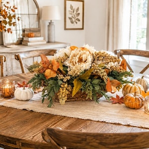 May include: A centerpiece arrangement featuring cream-colored peonies, golden yellow foliage, and green leaves housed in a woven basket. The display is surrounded by small white and orange pumpkins, scattered autumn leaves, and glass candle holders.