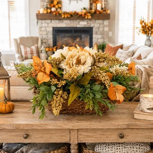May include: A rectangular woven basket filled with artificial cream-colored peonies, orange leaves, and golden-yellow sprigs. The arrangement sits on a wooden table alongside a metal lantern, two small pumpkins, a birch-style candle, and a sign reading grateful.