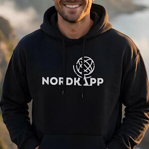 Puede incluir: Una sudadera negra con capucha que presenta un estampado gráfico blanco en el pecho. El diseño incluye el texto NORDKAPP en letras mayúsculas junto a un icono de globo terráqueo estilizado. La prenda tiene capucha con cordón y mangas largas.