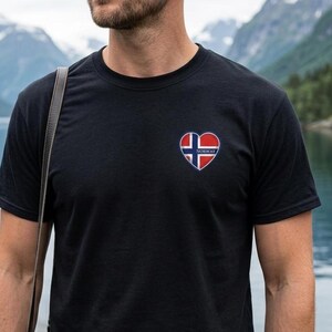 May include: A black short-sleeved t-shirt featuring a heart-shaped graphic on the left chest. The heart contains the red, white, and blue flag of Norway with the word NORWAY printed in white capital letters across the center of the design.