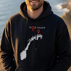 Op de afbeelding: Een zwarte hoodie met een witte afbeelding van de kaart van Noorwegen op de borst. Boven de kaart staat de tekst NORDKAPP in het wit gedrukt, met een rood cirkelvormig icoon en een rode locatiepin geplaatst op het noordelijke puntje.