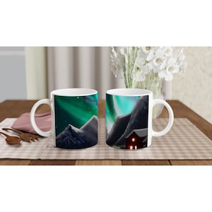 Puede incluir: Dos tazas de cerámica blanca con un diseño de una cordillera con una casa en primer plano y una aurora boreal verde y azul en el fondo.
