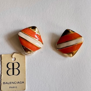 Puede incluir: Un par de pendientes dorados en forma de rombo, con rayas diagonales de esmalte naranja y blanco. Se adjunta una etiqueta rectangular de color blanco roto que muestra el logotipo negro de Balenciaga Paris y un símbolo de doble B estilizado.