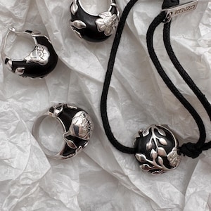 May include: A jewelry set consisting of two hoop earrings, a ring, and a black cord bracelet. Each piece features a smooth black material accented with polished silver-tone floral motifs. The bracelet includes a silver-tone tag labeled KENZO.