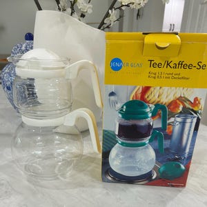 May include: A clear glass tea/coffee set with white and cream-colored handles and lids. The set includes a 1.5-liter carafe and a 0.5-liter carafe with a lid filter. The box reads "JENAER GLAS Tee/Kaffee-Set".