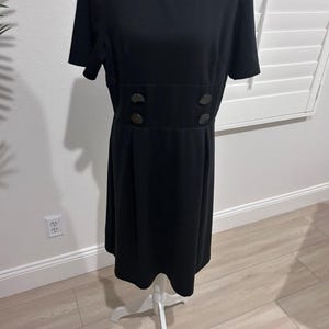 May include: A black dress with short sleeves and a round neckline. The dress features a fitted waist with four dark buttons and a flared skirt. The dress is displayed on a white mannequin.