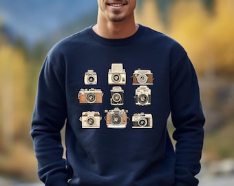 May include: Navy blue sweatshirt featuring a collection of vintage camera illustrations in various sizes and styles. The cameras are in shades of brown, beige, and black, creating a retro aesthetic. The sweatshirt is a comfortable, casual garment.