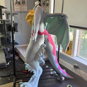 May include: A light blue and pink dragon fursuit with yellow accents, wings, and large feet. The costume is hanging on a clothing rack. The dragon's tail has a pink stripe. The costume is made of soft, plush fabric.