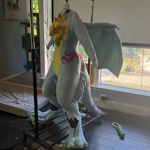 May include: A light blue and yellow dragon costume with large wings, hanging on a black clothing rack. The costume has pink accents and large feet. The costume is made of soft fabric.