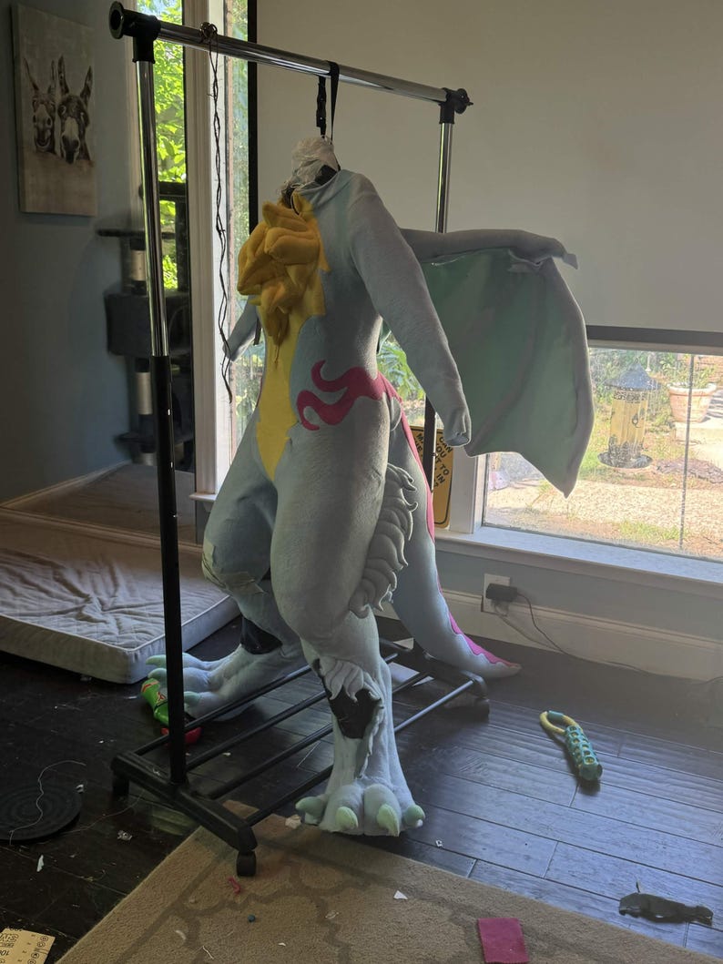 May include: A light blue and yellow dragon costume with large wings, hanging on a black clothing rack. The costume has pink accents and large feet. The costume is made of soft fabric.
