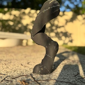 May include: A black, abstract sculpture with a curved, organic shape. The sculpture appears to be made of fabric or paper, with visible stitching and markings. It stands on a textured, light-colored surface, with a blurred background of greenery and a wall.