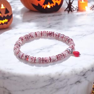 May include: A beaded bracelet with a Halloween theme. The bracelet features round, patterned beads in shades of red and white, interspersed with silver-colored spacers. A red charm hangs from the bracelet. The background includes carved pumpkins and candles.