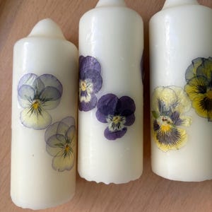 May include: Three white pillar candles with pressed flowers. The candles have purple and yellow flowers pressed into the wax.