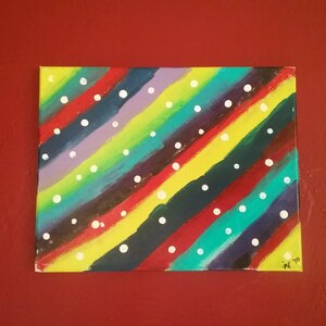 Könnte beinhalten: Ein farbenfrohes abstraktes Gemälde auf Leinwand. Diagonale Streifen in verschiedenen Farben, darunter Rot, Gelb, Grün und Lila, werden durch weiße Punkte akzentuiert. Das Kunstwerk ist an einer roten Wand befestigt.