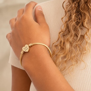 May include: A gold-toned cable wire bangle bracelet featuring a central heart-shaped charm. The charm is engraved with the text Emily and James separated by a small heart symbol. The bracelet has cylindrical metal accents on both sides of the charm.