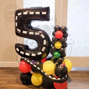 May include: A black number 5 balloon with white lines resembling a race track, surrounded by black, red, yellow, and green balloons. The balloons are arranged to resemble a traffic light and a race track.