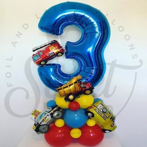 May include: A blue number 3 balloon with a red fire engine, a yellow dumper truck, a black and white police car, and a yellow school bus attached to it. The balloons are arranged in a tower shape with red, yellow, and blue balloons.