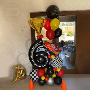 May include: A black number six balloon, a red race car balloon, a gold trophy balloon, and a variety of black, red, yellow, and white balloons. The balloons are arranged in a cluster and are tied together with string. The balloons are on a white surface.