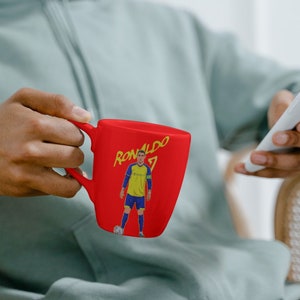 Puede incluir: Una taza de cer&aacute;mica roja con un gr&aacute;fico de un jugador de f&uacute;tbol y el texto "RONALDO 7".