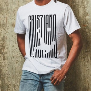 May include: White t-shirt with a black and white graphic print of the word "Cristiano" with a stylized image of a soccer player.