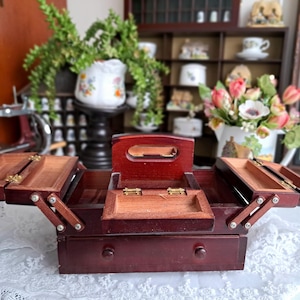 May include: A dark wood sewing box with a tiered, cantilevered design featuring multiple hinged compartments that fold outward. The box includes a central handle, a small interior tray, and a single bottom drawer with two small wooden knobs.