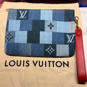 Op de afbeelding: Een clutch in denim-stijl met een patchwork-ontwerp in blauw- en grijstinten. De clutch heeft het Louis Vuitton-logo en een rode leren polsband. De tas ligt op een beige Louis Vuitton stofzak.