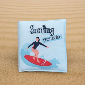 May include: A light blue baby book with the words "Surfing For Babies" and an illustration of a person surfing on a red surfboard. The book is on a sandy surface.