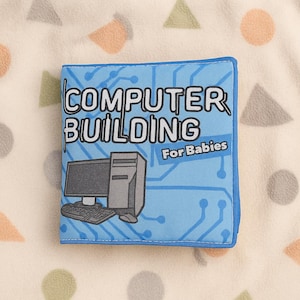 May include: A blue soft book titled "COMPUTER BUILDING For Babies" with a computer graphic. The cover features a circuit board design and illustrations of a computer monitor, keyboard, and tower. The book is on a cream-coloured surface with colourful shapes.