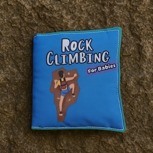 May include: A blue fabric book with the words "Rock Climbing For Babies" and an illustration of a person climbing a rock face. The book has a teal border and is on a textured brown surface.