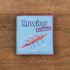 May include: A children's book titled "Rowing For Babies" with a light blue cover and pink trim. The cover features an illustration of a red rowboat with four people rowing. The book is set against a wooden background.