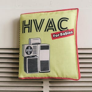 May include: A square, light green pillow with a red border. The pillow features a graphic of an HVAC unit and the text "HVAC For Babies" in black and red. The pillow is resting against a white surface with horizontal lines.