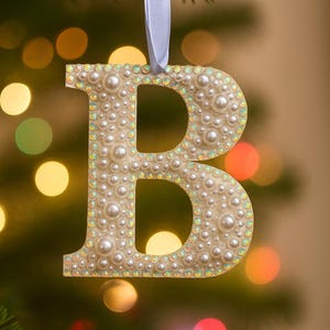 May include: A decorative Christmas ornament shaped like the letter "B". The ornament is covered in small, white pearls and iridescent rhinestones. It hangs from a light blue ribbon. The background is blurred with colorful lights.