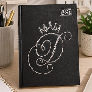 Könnte beinhalten: Ein schwarzer 2027 Tagesplaner mit einem großen, kursiven Buchstaben D, der mit klaren Strasssteinen verziert ist. Über dem Buchstaben befindet sich ein Strass-Kronendesign. Der Einband ist texturiert und der Text 2027 Day A Page ist weiß gedruckt.